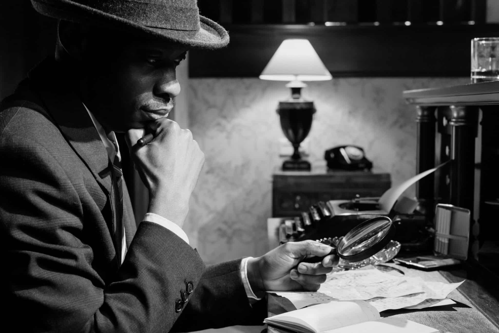 A man looking at a suspicious notebook using a magnifying glass with his hand under his chin wearing a hat. 