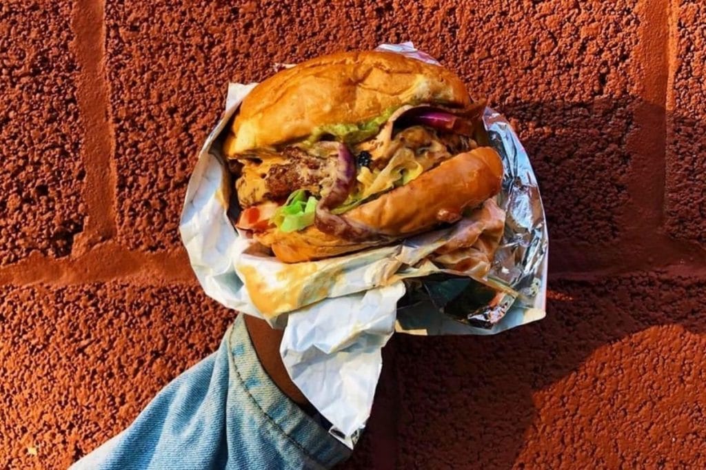 A close up of a hand holding a big vegan burger against a red-brick wall