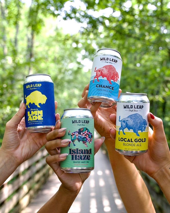 An image of four hands holding up different wild leap beer cans with a green tree background 