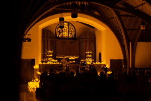 The silhouette of an audience sitting before an altar lit up by the glow of candlelight while musicians perform a Candlelight concert.