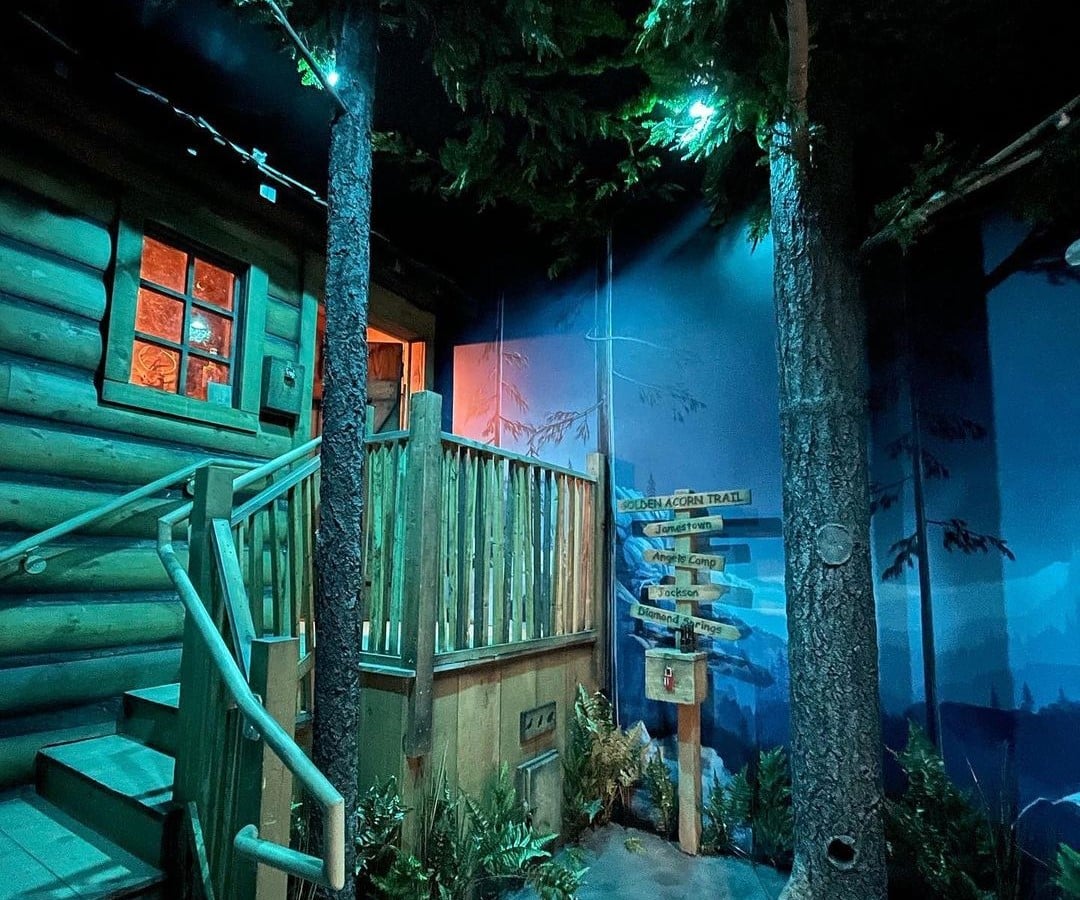 An escape room with a wooden cabin, trees, and sign posts set up. 