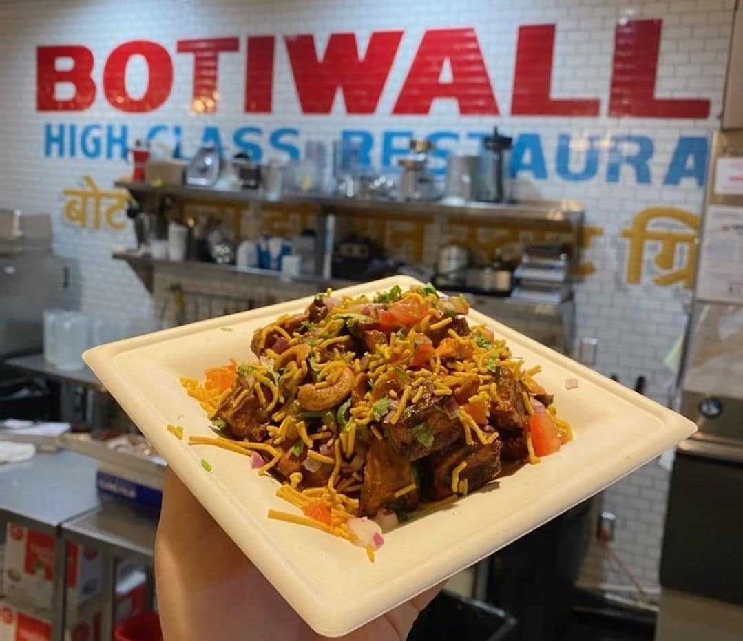A plate of Indian food being held up to the camera with "Botiwalli" written on the kitchen wall.