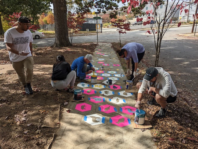 A photo of volunteers painting symbols of the Muscogee Nation on tiles on a sidewalk. Mulch and Trees in the background.