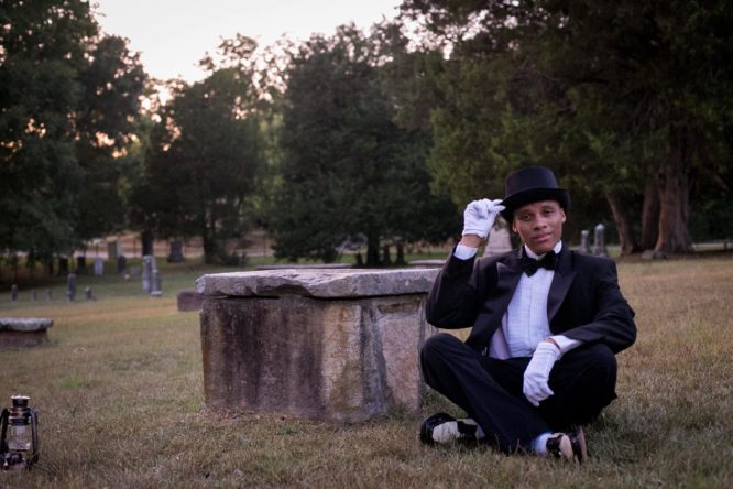 man sitting in victorian clothes near a graveyard