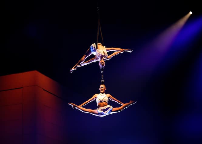 two women acrobats, hanging in full splits by their hair, which is in a bun